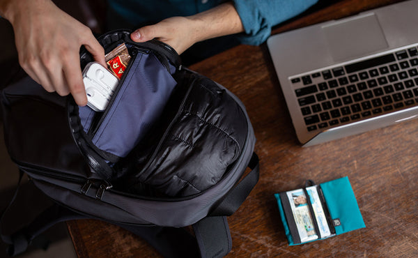 A person removing some earbuds and a protein bar from the top interior zippered pocket, also showing a jacket held in the main pocket.